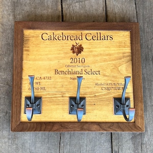 May include: A wooden wine rack with three hooks, featuring a label that reads "Cakebread Cellars 2010 Cabernet Sauvignon Benchland Select Napa Valley Alcohol 14.5% By Volume CSB0710W".
