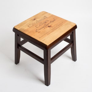 May include: A dark brown wooden stool with a square top. The top is made of wood with a light brown stain and has the text "Cakebread Cellars Dancing Bear Ranch 2012 Grown, Produced & Bottled by Cakebread Rutherford, California, USA" printed on it.