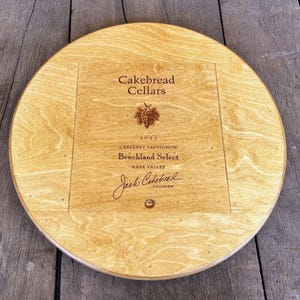 May include: A round wooden wine barrel top with a label that reads "Cakebread Cellars 2021 Cabernet Sauvignon Benchland Select Napa Valley Jack Cakebread Founder".