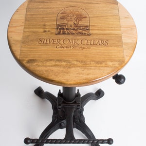 May include: A wooden bar stool with a round, brown wood top featuring a laser-engraved logo that reads "Silver Oak Cellars Cabernet Sauvignon". The stool has a black metal base with a footrest.