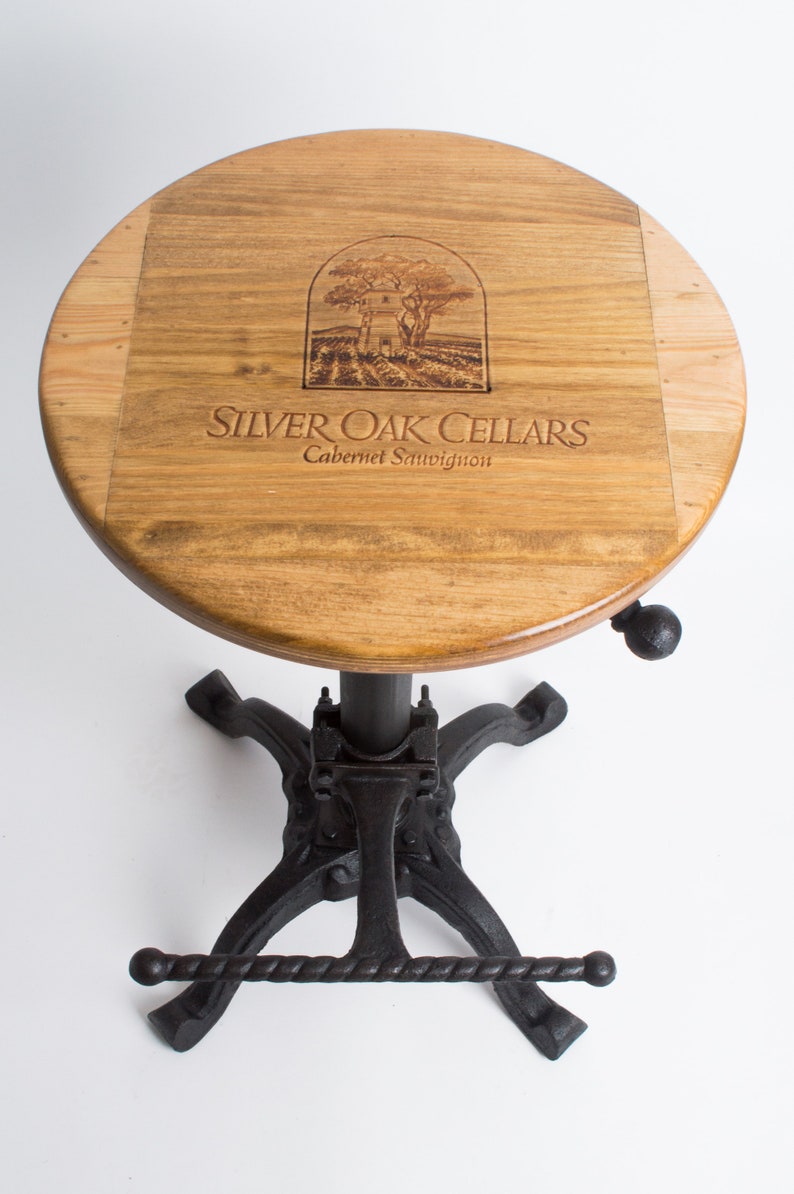 May include: A wooden bar stool with a round, brown wood top featuring a laser-engraved logo that reads "Silver Oak Cellars Cabernet Sauvignon". The stool has a black metal base with a footrest.