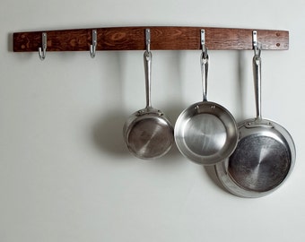 May include: A wall-mounted kitchen utensil rack made from a reclaimed oak barrel stave. The rack has five hooks and is holding three stainless steel pots.