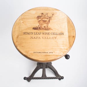 May include: A round wooden table top with a stag's leap wine cellars logo, featuring a deer and trees, and the text "Napa Valley" and "Established 1970". The table has a black metal base with a footrest.