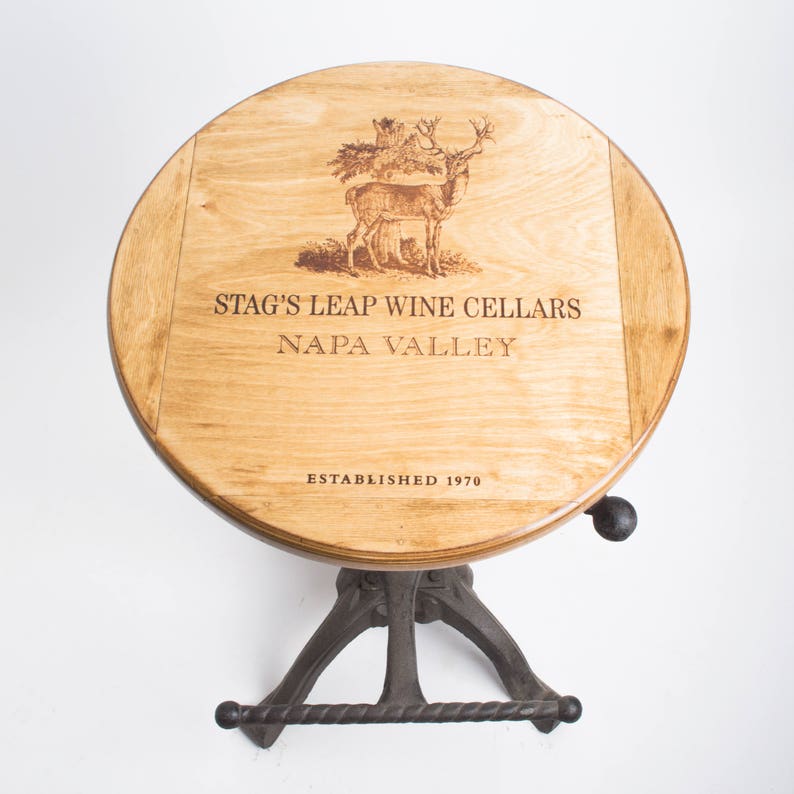 May include: A round wooden table top with a stag's leap wine cellars logo, featuring a deer and trees, and the text "Napa Valley" and "Established 1970". The table has a black metal base with a footrest.