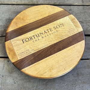 May include: A round wooden serving tray featuring alternating light and dark wood grain panels. The center is laser-engraved with the text FORTUNATE SON THE WARRIOR, along with details including 3x750ml 2.25ml, 15.5% alc. by volume, and Napa Valley California.