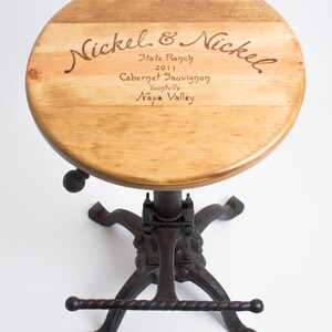 May include: A wooden bar stool with a round, wooden seat featuring an engraved wine label that reads "Nickel & Nickel, State Ranch, 2011, Cabernet Sauvignon, Yountville, Napa Valley". The stool has a black metal base with a footrest.