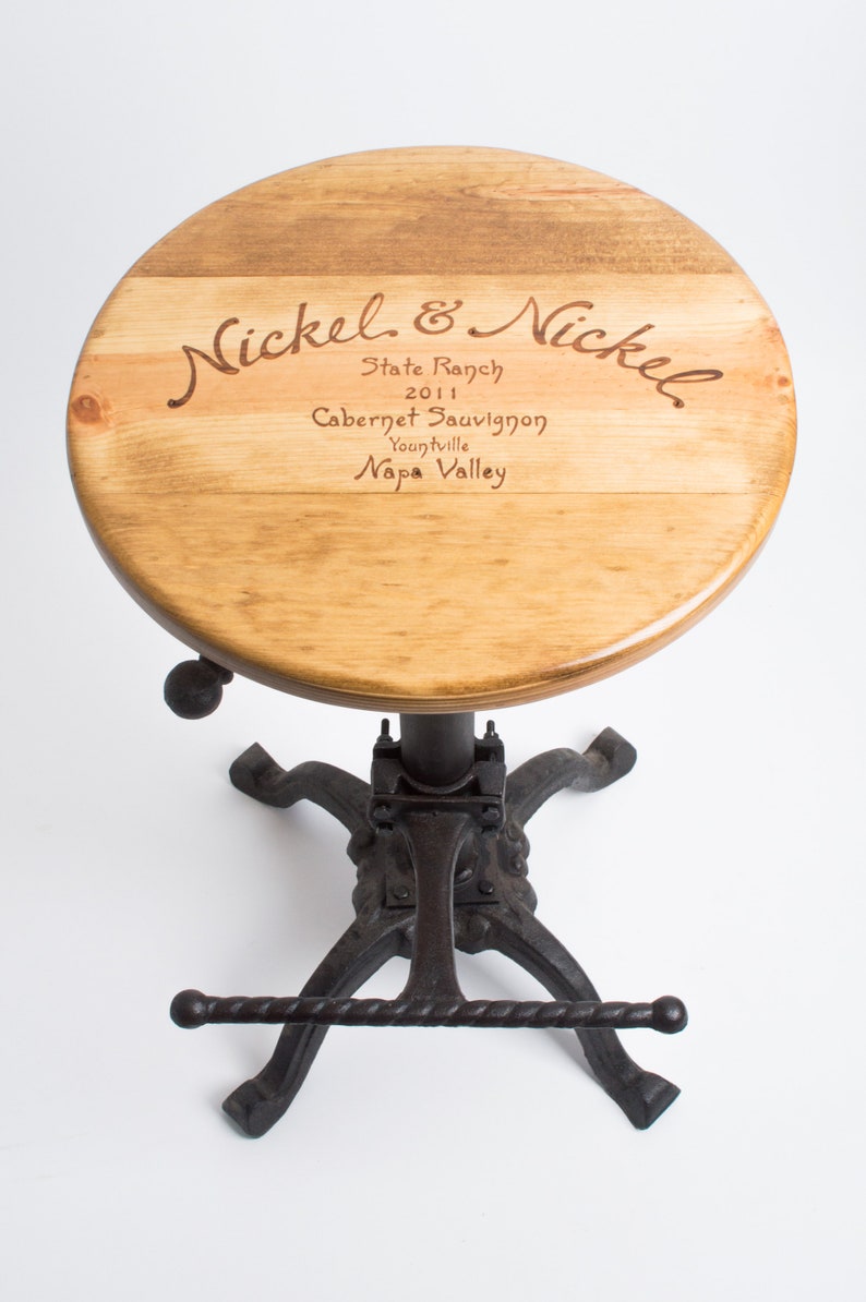 May include: A wooden bar stool with a round, wooden seat featuring an engraved wine label that reads "Nickel & Nickel, State Ranch, 2011, Cabernet Sauvignon, Yountville, Napa Valley". The stool has a black metal base with a footrest.