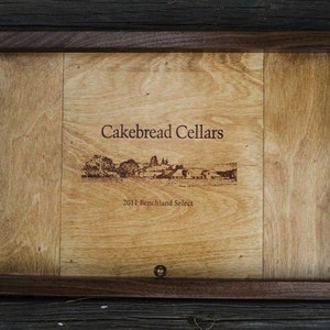 May include: A wooden serving tray with a dark brown frame and a light wood interior. The tray has a laser-etched design of a vineyard and the text "Cakebread Cellars 2011 Benchland Select".