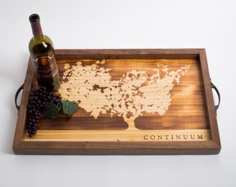 May include: A wooden serving tray with a burnt-in design of a tree and the word "CONTINUUM". The tray has two metal handles on the sides. A bottle of wine and grapes are on the tray.