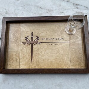 May include: A rectangular wooden serving tray with dark brown edges and a lighter wood center panel. The tray features a dark brown laser-engraved design of a sword and crown with the text "FORTUNATE SON" and "THE WARRIOR" and "18 69". A clear glass is placed on the tray.