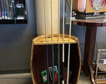 May include: A wooden barrel repurposed as a floor-standing pool cue rack. The rack features a circular top with five holes for holding cues vertically. Five pool cues are inserted into the rack, displaying various colors including blue, green, and white.