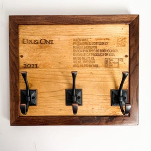 May include: A wooden coat rack with three hooks, made from a repurposed wine bottle label. The label features the text "Opus One" and "2021". The rack is framed in dark wood.