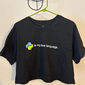 May include: Black crop top with the text "is my love language." The shirt features a Python logo in yellow and blue. The shirt is hanging on a green hanger.