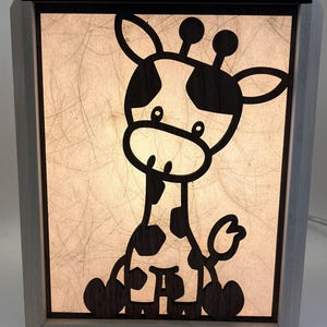 May include: A wooden night light with a giraffe silhouette. The giraffe is black with white spots and is set against a cream-colored background. The night light has a rectangular shape with a light wood frame and a black top.