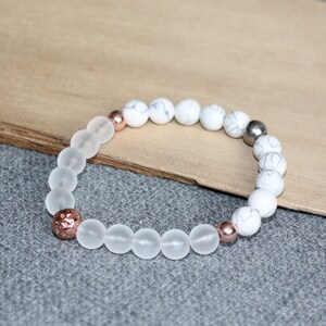 May include: A beaded bracelet featuring frosted glass and white marble-like beads. Accented with rose gold and silver-colored beads. The bracelet is displayed on a wooden surface against a gray background.