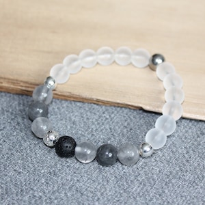 May include: A beaded bracelet featuring frosted white, gray, and black beads. The bracelet includes silver-colored metal accents and a black lava bead. The beads are strung on an elastic band.