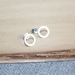 May include: A pair of small, silver-colored circle stud earrings. The earrings are simple in design, featuring a thin, open circle shape. They are displayed on a light brown wooden surface.