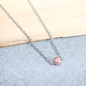 May include: A delicate silver necklace featuring a small, rose-gold colored bead pendant. The chain has a fine, textured design. The pendant is a small, round bead with a textured surface. The necklace is displayed on a light gray surface.