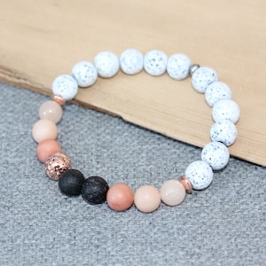 May include: A beaded bracelet featuring a mix of white speckled, black, and peach-colored round beads. The bracelet includes rose gold-colored accents and is displayed on a neutral gray surface.