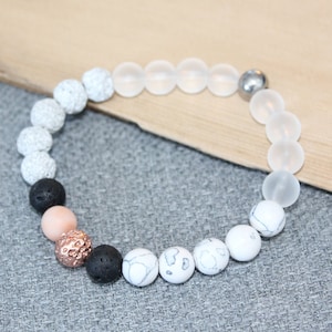 May include: A beaded bracelet featuring a variety of stone beads in shades of white, gray, black, and rose gold. The bracelet includes lava stone, matte white, and textured beads, with a silver-colored clasp. The bracelet is displayed on a gray surface.