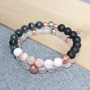 May include: Two beaded bracelets with a variety of stone beads. One bracelet features black lava beads, rose quartz, and a rose gold accent bead. The other bracelet has gray, white, and pink beads with rose gold accents. The bracelets are on a wooden surface.