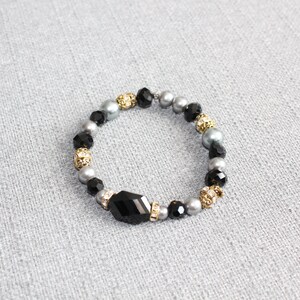 May include: A stylish bracelet with a combination of black, grey, and gold beads. The bracelet incorporates faceted black beads, round grey beads, and gold-coloured, rhinestone-studded beads. A large, black, faceted centrepiece is included.