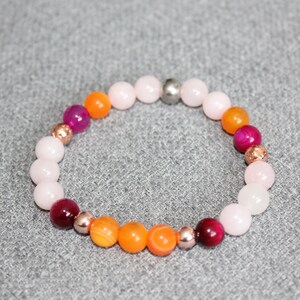 May include: A beaded bracelet featuring a mix of round beads in shades of pink, orange, and burgundy. Accents include metallic beads. The bracelet is displayed on a gray surface.