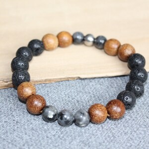 May include: A beaded bracelet featuring a mix of natural materials. The bracelet includes black lava rock beads, light brown wooden beads, and metallic gray beads. The beads are strung together to form a circular shape.