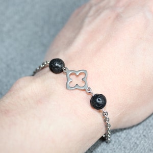 May include: A silver-toned bracelet featuring a clover-shaped charm and two black lava stone beads. The bracelet is made of a delicate chain and is shown against a gray background. The jewelry is a simple, elegant accessory.