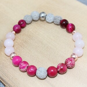 May include: A beaded bracelet featuring a mix of pink, red, and gray stones. The bracelet includes rose gold accents and is displayed on a wooden surface. The beads vary in size and texture, creating a visually appealing accessory.