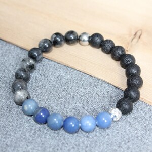 May include: A beaded bracelet featuring a mix of natural stone beads. The bracelet includes blue, gray, and black lava stone beads, with a silver accent bead. The bracelet is displayed on a gray surface with a wooden background.