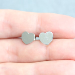 May include: A pair of small, silver-colored heart-shaped stud earrings. The earrings are displayed against a light blue background, held by a person's fingers.