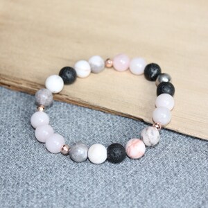 May include: A beaded bracelet featuring a mix of round beads in various colors and textures. The bracelet includes pink, white, gray, and black beads, with a few beads having a speckled appearance. The beads are strung together with rose gold-colored spacers.