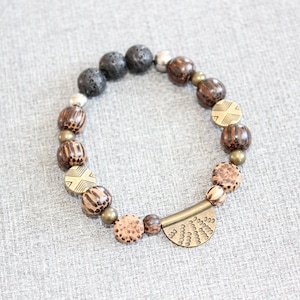 May include: A beaded bracelet featuring a mix of dark lava stone, silver, and wooden beads. The bracelet includes patterned wooden beads and a decorative brass-toned half-moon accent. The bracelet is on a gray surface.