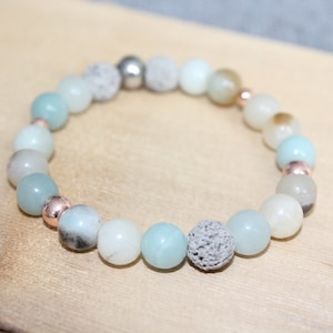 May include: A beaded bracelet featuring a mix of light blue, white, and gray round beads. The bracelet includes rose gold-colored spacers and a textured gray bead. The bracelet is displayed on a light wood surface.