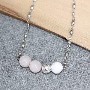 May include: A delicate necklace featuring a silver chain with three light pink beads, a textured silver bead, and a white bead. The necklace is displayed on a grey surface with a wooden accent in the background.