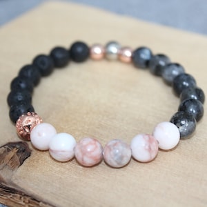 May include: A beaded bracelet featuring a mix of natural stone beads. The bracelet includes black lava beads, pink and white patterned beads, and metallic accents. The beads are strung together on an elastic cord, creating a stretchable design.