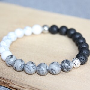 May include: A beaded bracelet featuring a mix of white, gray, and black beads. The bracelet has a silver accent bead and is displayed on a wooden surface. The beads have a variety of textures and patterns.