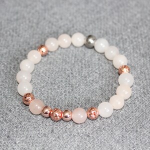 May include: A beaded bracelet featuring a mix of pale pink and white round beads. Accented with rose gold-colored, textured beads and a single silver-colored bead. The bracelet is displayed on a gray surface.