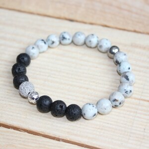 May include: A beaded bracelet featuring a mix of black lava rock, speckled white, and silver-toned beads. The bracelet is strung on an elastic cord and is displayed on a light wood surface. This accessory is suitable for everyday wear.