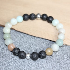 May include: A beaded bracelet featuring a mix of colors and textures. The bracelet includes round beads in shades of white, green, and tan, interspersed with black lava beads and a silver-colored bead. The bracelet is displayed on a gray surface.