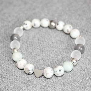 May include: A beaded bracelet featuring a mix of white, gray, and silver beads. The bracelet includes speckled white beads, clear and gray round beads, and a silver heart charm. The bracelet is displayed on a gray surface.