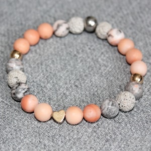 May include: A beaded bracelet featuring peach-colored, gray, and gold-colored beads. The bracelet includes a small gold heart charm. The beads are arranged in a circular pattern.