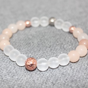 May include: A beaded bracelet featuring a mix of light pink and frosted white beads. Accents include rose gold and silver-colored beads. The bracelet is designed with a stretchable band.