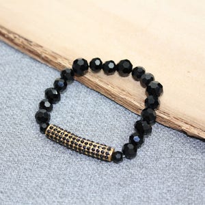 May include: Black beaded bracelet with a gold-colored, rhinestone-studded bar. The bracelet features faceted, round black beads. The gold bar is centered on the bracelet. The bracelet is displayed on a gray surface with a wooden accent.