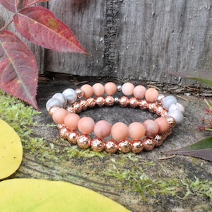 May include: Two stacked bracelets with peach-colored and silver beads. The bottom bracelet has textured, rose gold-colored beads. The bracelets are displayed on a mossy, wooden surface with colorful fall leaves.