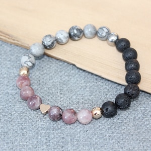 May include: Beaded bracelet featuring a mix of natural stone beads. The bracelet includes pink, gray, and black lava stone beads, along with gold-colored accents. A small gold heart charm adds a decorative touch to the bracelet.