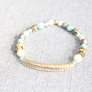 May include: A beaded bracelet featuring a gold-tone curved bar with embedded rhinestones. The bracelet includes turquoise, white, silver, and gold beads. Decorative metal accents are interspersed throughout the design. The bracelet is on a gray surface.