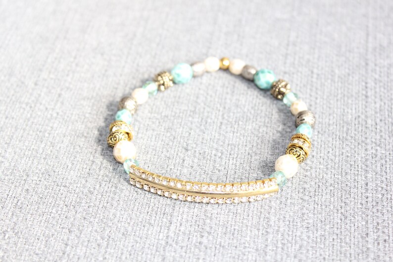 May include: A beaded bracelet featuring a gold-tone curved bar with embedded rhinestones. The bracelet includes turquoise, white, silver, and gold beads. Decorative metal accents are interspersed throughout the design. The bracelet is on a gray surface.