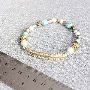 May include: A beaded bracelet with a gold-tone bar embellished with small, clear rhinestones. The bracelet features a mix of white, turquoise, silver, and gold beads. The bracelet is approximately 7 inches in length.